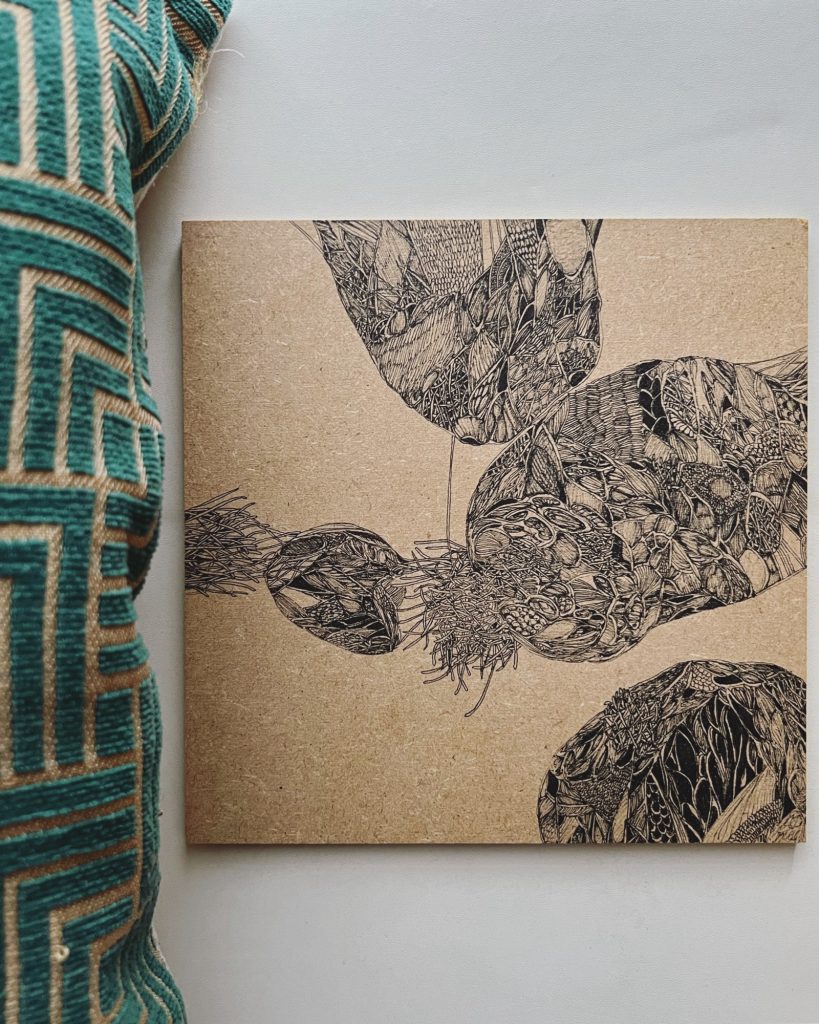 Dessin fin d’inspiration cellulaire et organique en noir, sur une plaque de bois carrée de 18 cm par 18 cm à côté d’un coussin vert à motifs dorés. 