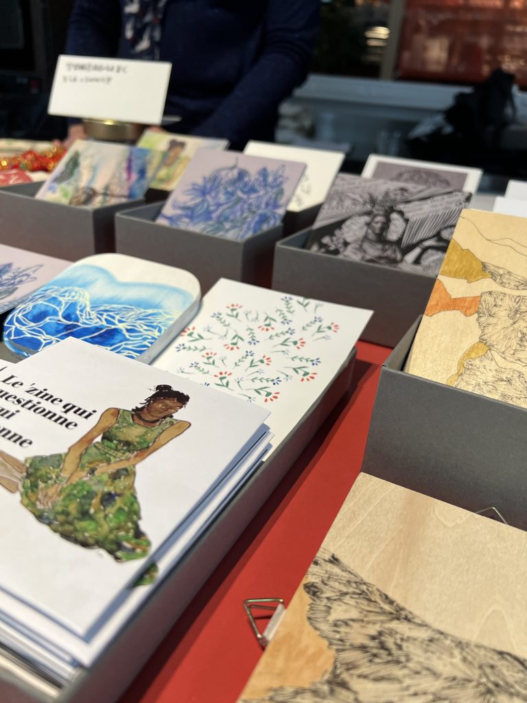 On voit une table d’exposition sur laquelle sont posés des œuvres : des fanzines en papier, des illustrations en série limitée et des dessins sur bois, les dessins sont fins en couleurs ou en noir et blanc. Les tons de couleurs entre le bleu le vert et le gris. La nappe de la table est rouge car c’était pendant le nouvel an chinois. 