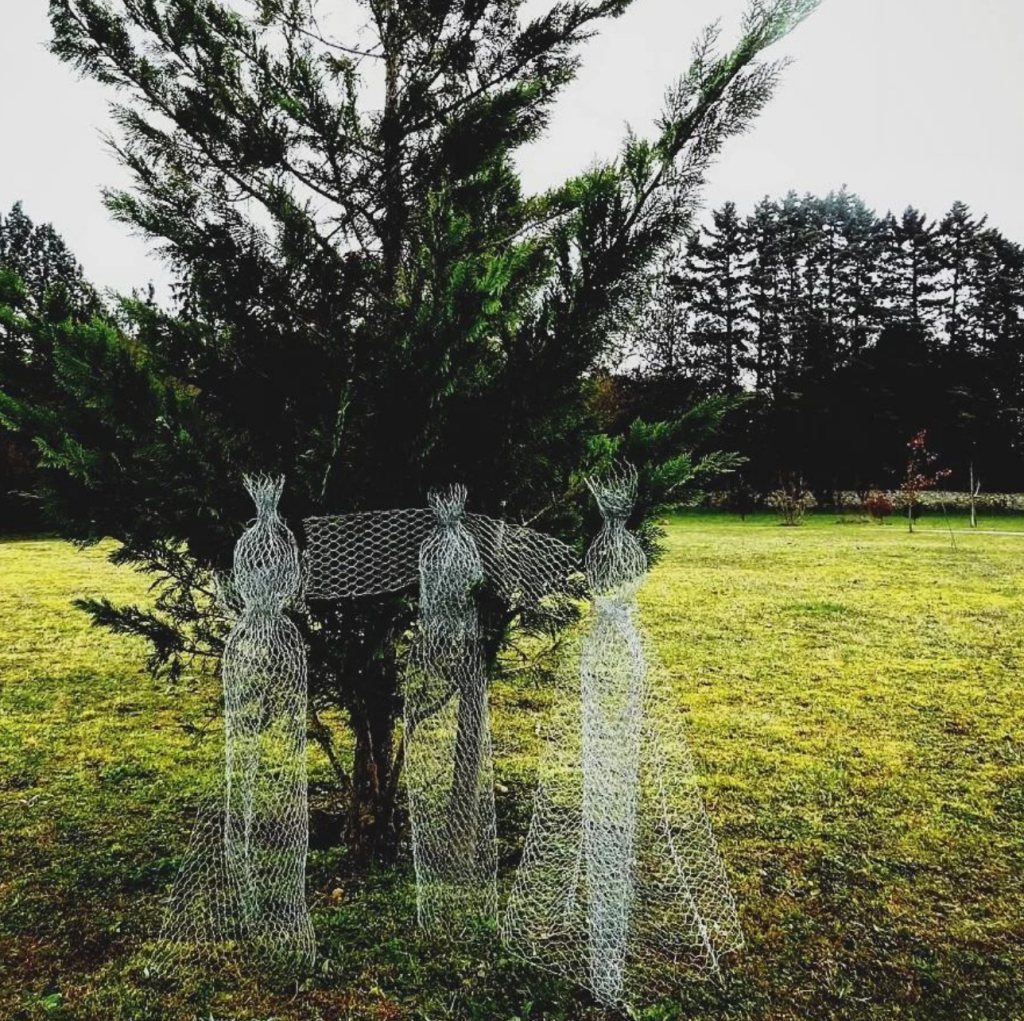 sculpture en grillage dans une position "bras ouverts" pas de tête devant un thuya