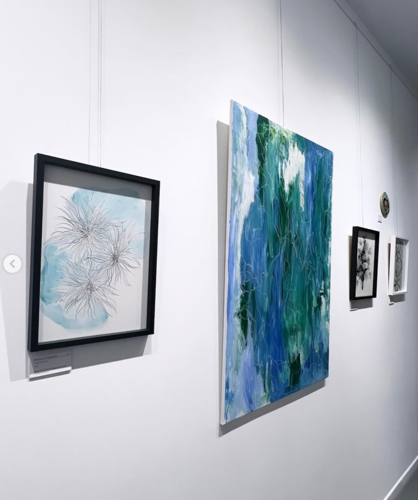 Photographie de côté de l'exposition avec un dessin aquarelle bleu et motifs végétaux en noir, et a côté une toile peinture acrylique abstraction vert bleu blanc plus loin deux autres dessins, encadrement noir et blanc